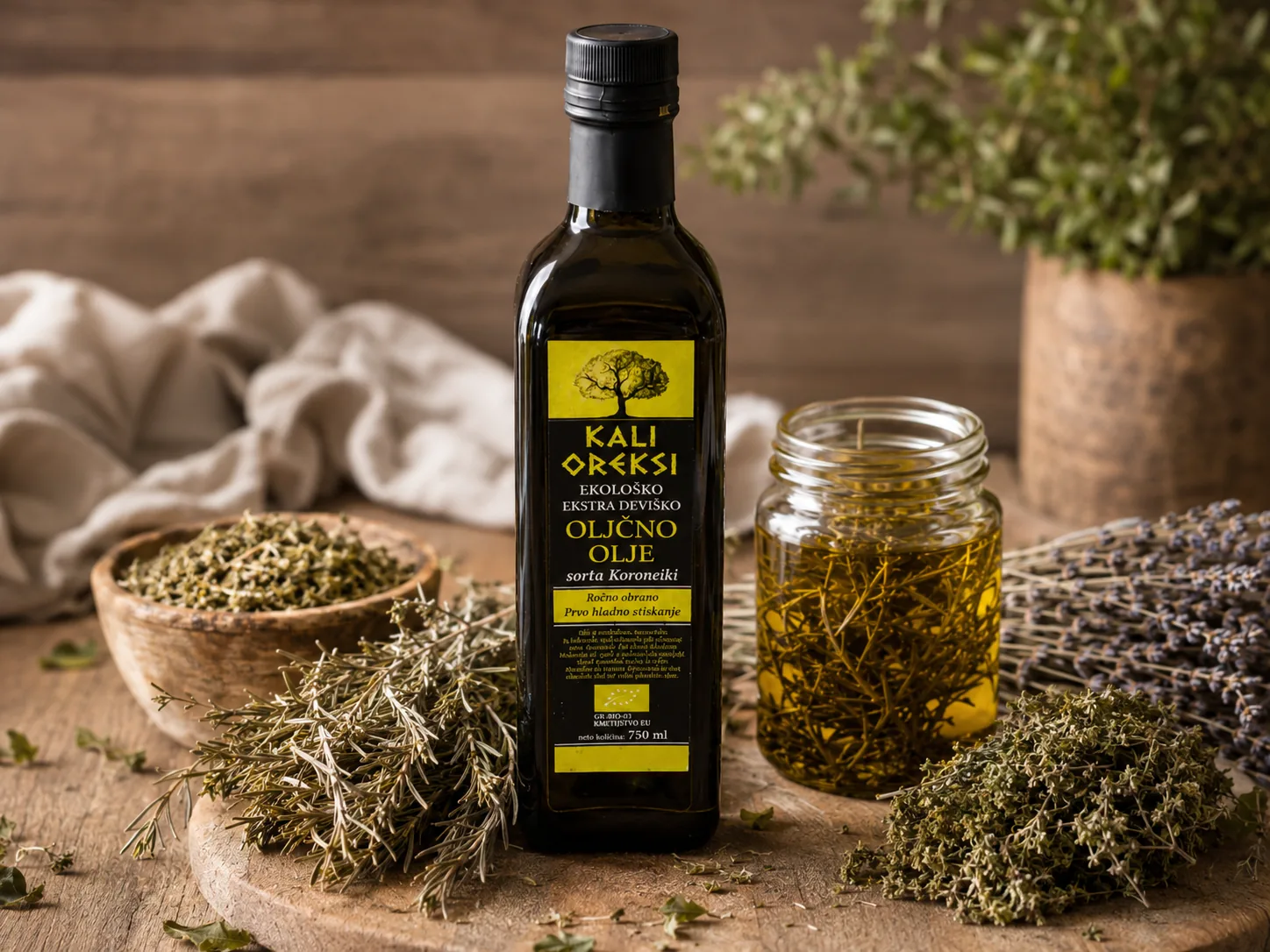 Steklenica oljčnega olja Kali Oreksi in kozarec olja z vejicami rožmarina, ob njem suh origano in sivka na leseni mizi